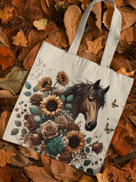 Canvas tote bag with boho style chocolate brown horse, surrounded by sunflowers, roses, and other flowers in blue, brown, and yellow hues. Tote is laying on a bed of autumn leaves.