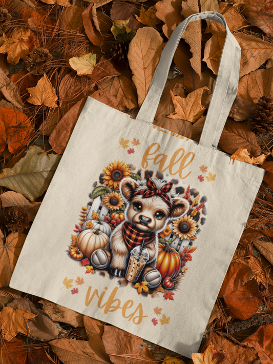 Canvas Tote Bag printed on both sides with cute highland cow wearing orange and black plaid headband and bandana. Cow is holding iced coffee, surrounded by pumpkins fall foliage, and sunflowers, on a cheetah print background. Text in light orange reads "fall vibes". Tote is laying on a background of autumn leaves.