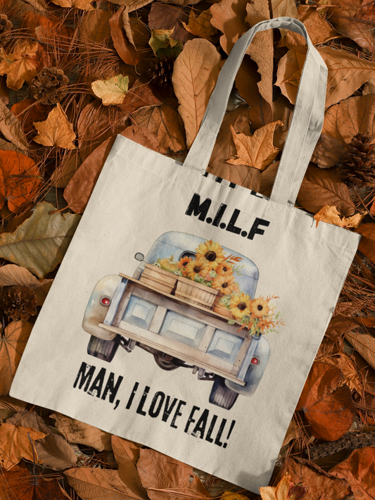 Canvas tote with the image of the back of a classic truck carrying whiskey barrels filled with sunflowers. Text above image reads "MILF". Text below image reads "Man, I love fall". Tote is laying on a bed of autumn leaves.