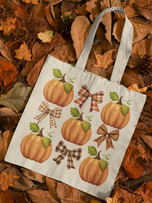 Trendy canvas tote bag with a plaid coquette bows and pumpkins in an alternating 9-space grid design. Bag is laying on a bed of autumn leaves.